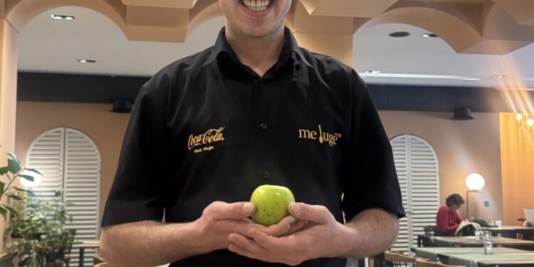 Jeton Musa holding a green apple at Meluge, with Chancellor Speedy praising his service as part of The Owner Schools' grassroots approach to supporting small businesses.