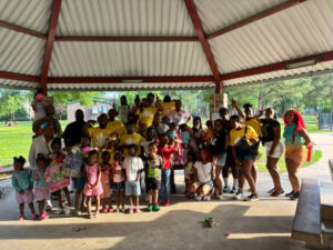 A large group of smiling event participants, including children and adults, pose together outdoors during KWAM Tha Barber’s Easter community event, surrounded by colorful decorations and festive energy.