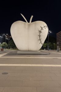 A large white apple sculpture with visible steel stitching, situated in front of Milan’s Central Station
