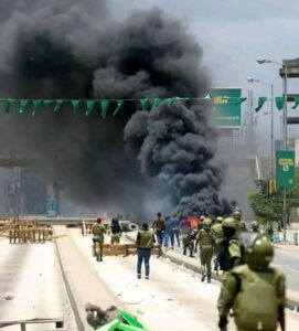 A crowd of Tanzanian protesters gathered on a city street at dusk. Small fires burn on the road, and smoke rises as people stand and watch. The scene conveys public unrest and civic tension.