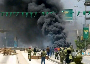 A crowd of Tanzanian protesters gathered on a city street at dusk. Small fires burn on the road, and smoke rises as people stand and watch. The scene conveys public unrest and civic tension.
