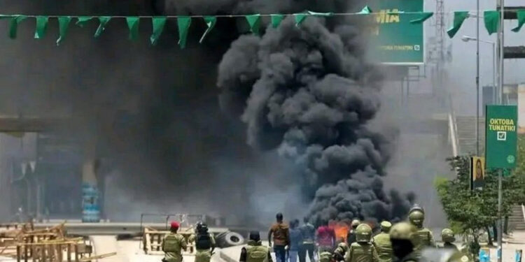A crowd of Tanzanian protesters gathered on a city street at dusk. Small fires burn on the road, and smoke rises as people stand and watch. The scene conveys public unrest and civic tension.